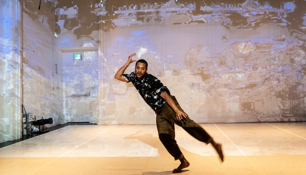 Styles Alexander a black man with close cropped hair and a mustache balances on one foot on a stage with projections behind him. He wears a black  and whit top and brown pants.