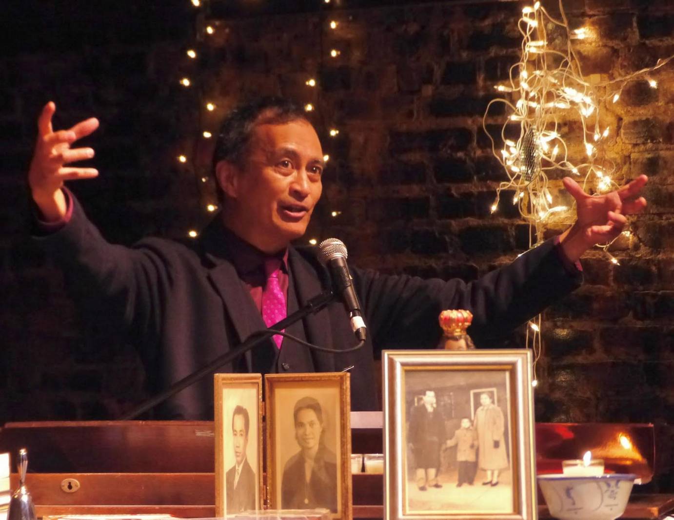 Nicky Paraiso in mid conversation arms extended outward, mic at his lips. He wears a black suit and shirt set off by a hot pink tie. Portraits of his parents from the Philippines sit on the dias.