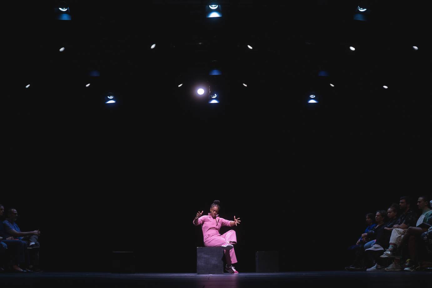 black dancer in a pink work uniform sits in the center of two groups of audience members. Her legs are crossed and her arms are open in front of her with fingers extended.