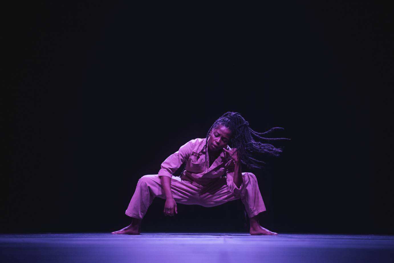 a black woman with long dreads faces us. Her legs are spread wide and bent in an almost balletic squat. one hand is to her ear as if she is talking on the phone