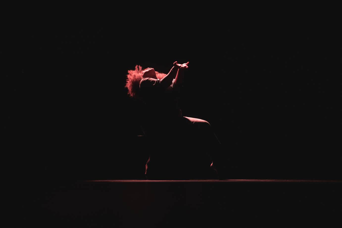 a light skinned black woman with a big reddish brown cirlce of hair, sits down on some sort of chair we cant see. Her face goes directly into the light and her bent arms and hands reach toward it