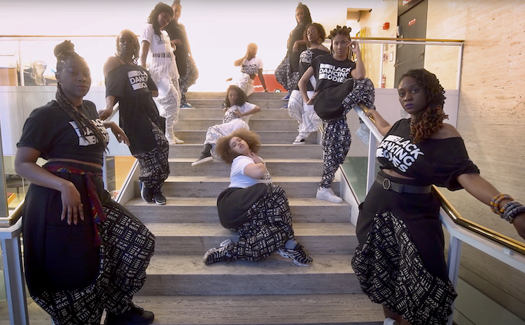 a company of black women in t-shirts and balloon-like pants in white of white print on black grace the stairs of the Lincoln Center Perfroming Arts Library. They stare out at us