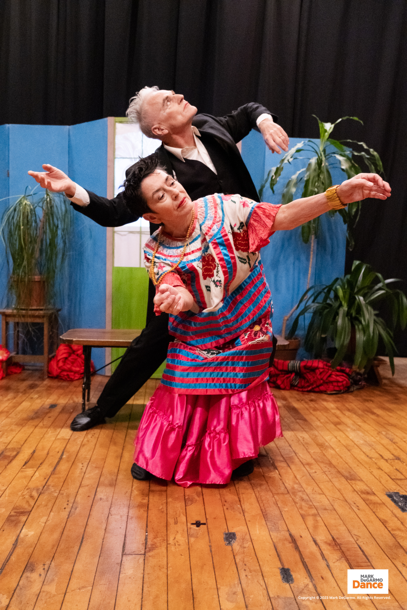 scenery representing Frida Kahlo's blue house is the back ground as a mane with grey hair and strong facial features looks upward, in front of him an older woman in a bright multi-colored Mexican dress bends forwards her arms extended in front of her, she seems to gaze inwardly, thoughtful.