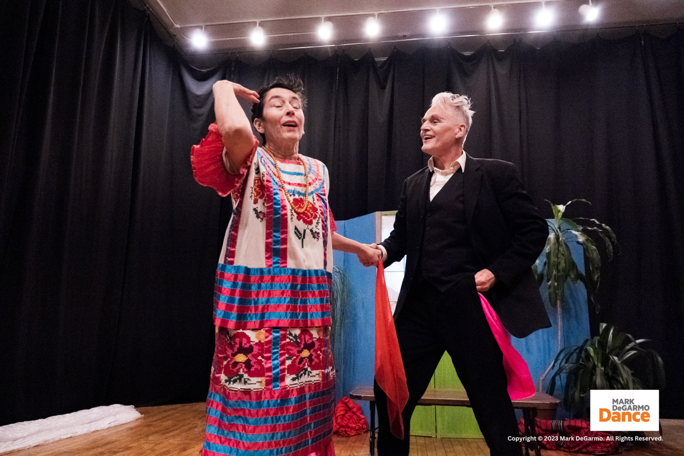 the woman in bright mexican dress holds hands with the man in a suit they both have bright red scarves in their hands as well