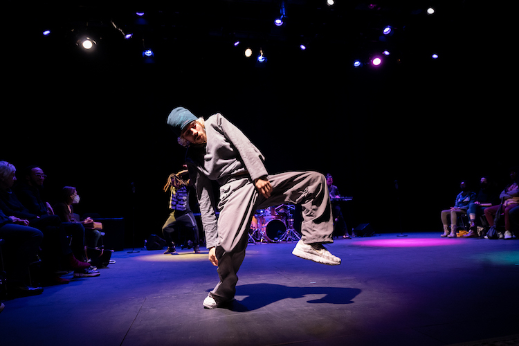 a fair skinned man with a blue cap , grey sweatshirt , grey pants,and sneakers dances in the center of a seated audience with castmates behind him. He is caught in the middle of a pose facing us with one hand brushing his chin and the other grabbing at his crotch as he stands on one leg. He is intense in concentration