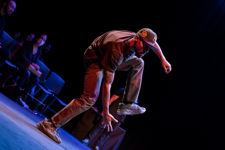 captured by the photographer from down below on an angle, a Black male soloist performs a solo with a masked audience member looking on. He reaches one arm to the floor balancing on one leg as he bends his torso forward as if to touch the floor.