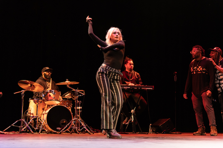 a fair-skinned woman with blonde hair , possibly from the audiences holds her arms up as if in a flamenco dance position as the cast looks on