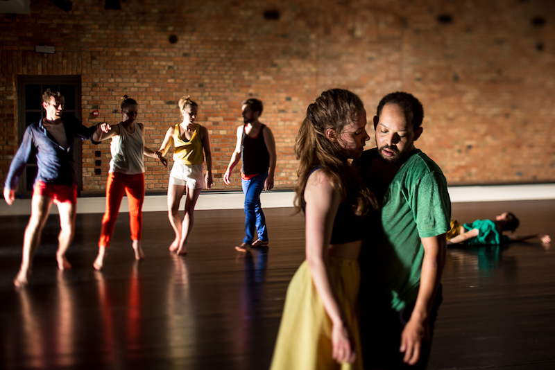 A man and woman stand chest to chest in the foreground while a line of dancers hold each others wrists while another dancer lays on the ground in the back