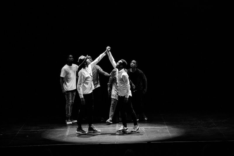 A black and white photo of two men hold one another's hand over their head. A group of performers huddle around them.