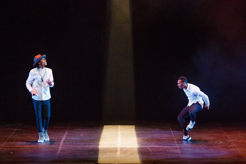 Two men in white collared shirts and jeans share the stage. A strip of light separates them.