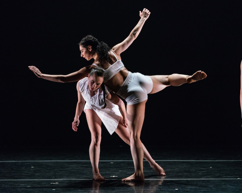 One woman leans her body over another as if she was using her counterpart's body as a table to rest on. They both wear white.