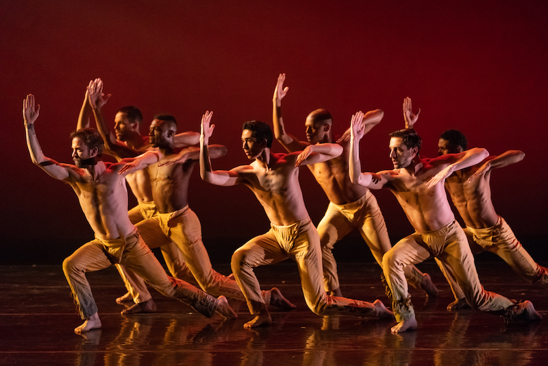 A group of men in tan pants stand in a lunge with their right arms raised.