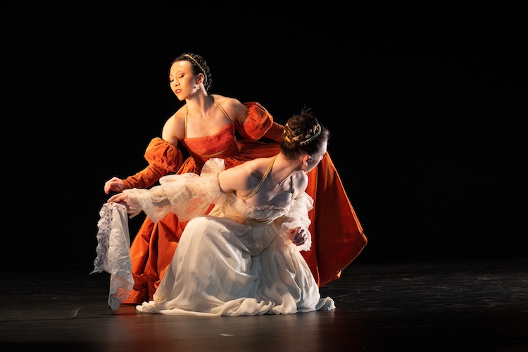 two women in courtly dress...one in orange, one in white in the middle of a dramatic scene.