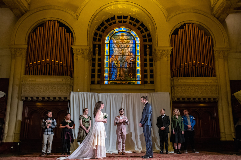 Two actors dress as bride and groom; others in casual dress stand as witnesses.