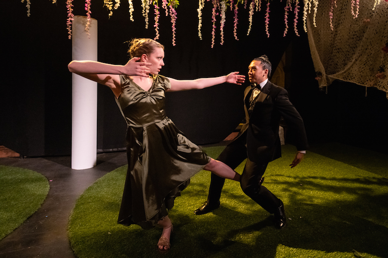 A man in a tuxedo dances with a woman wearing a sage sain evening gown. she positions her arms like she's pulling a bow and arrow. Her ankle hooks in his bent leg.