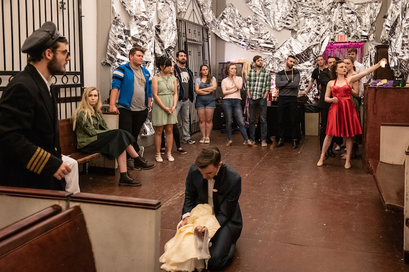 A man kneels on the ground clasping a white dress. A man in a navy coat and hat is in the foreground while audience members look at a woman in a red satin dress in the background.