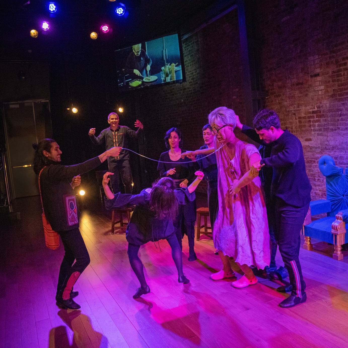 Sonia's oxygen tube is used for the limbo. Seen from behind a woman dressed in a blue dress bends low while others look on. A screen in the background features a chef preparing a meal.