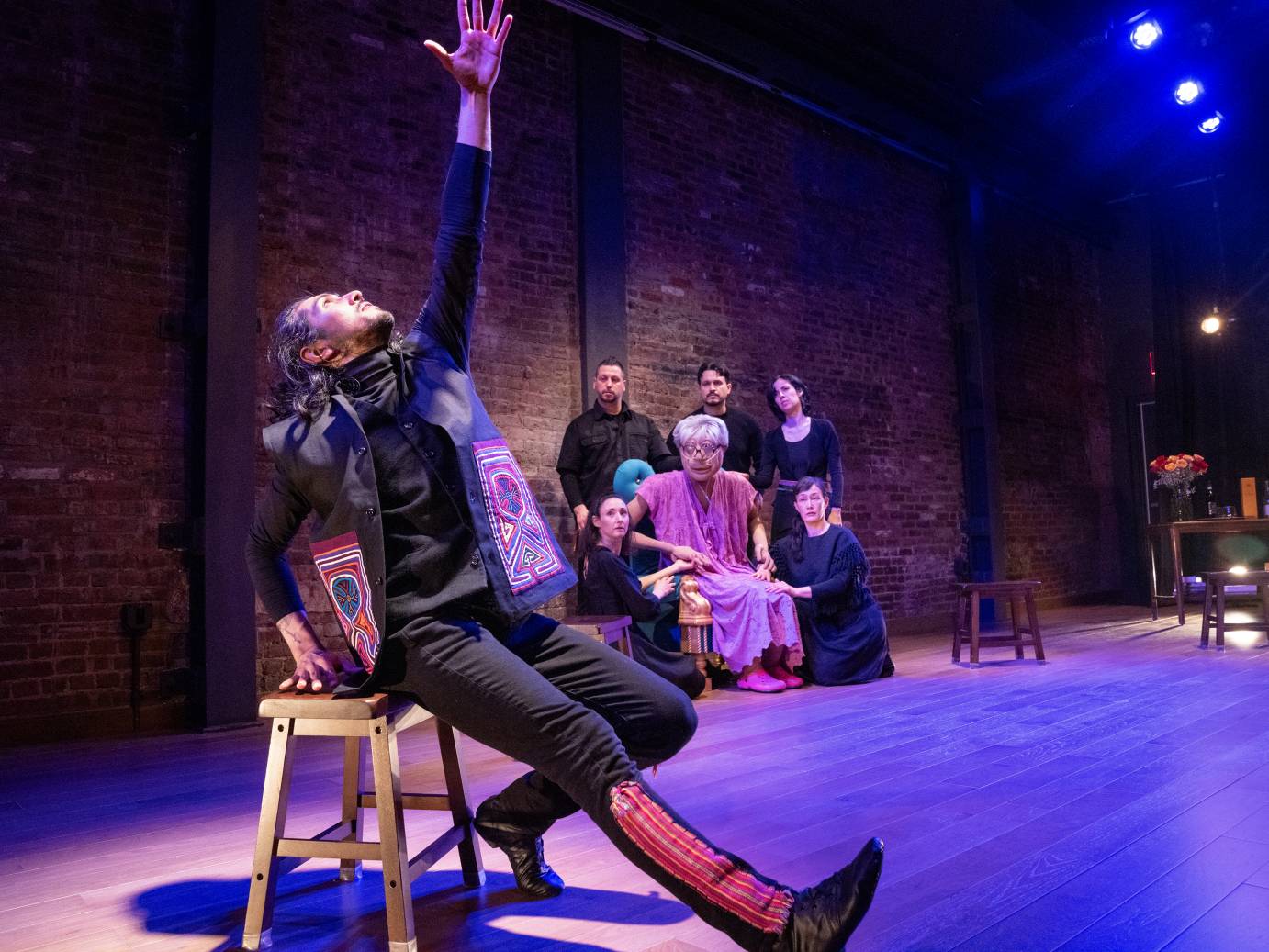 A dancer, seated on a blond wood stool, sports chin hair and hair to shoulders, wearing black clothing decorated with native Columbian designs. He reaches his open palmed hand to the ceiling while extending his right leg. He looks up. Puppet Sonia and five puppet handlers look on.