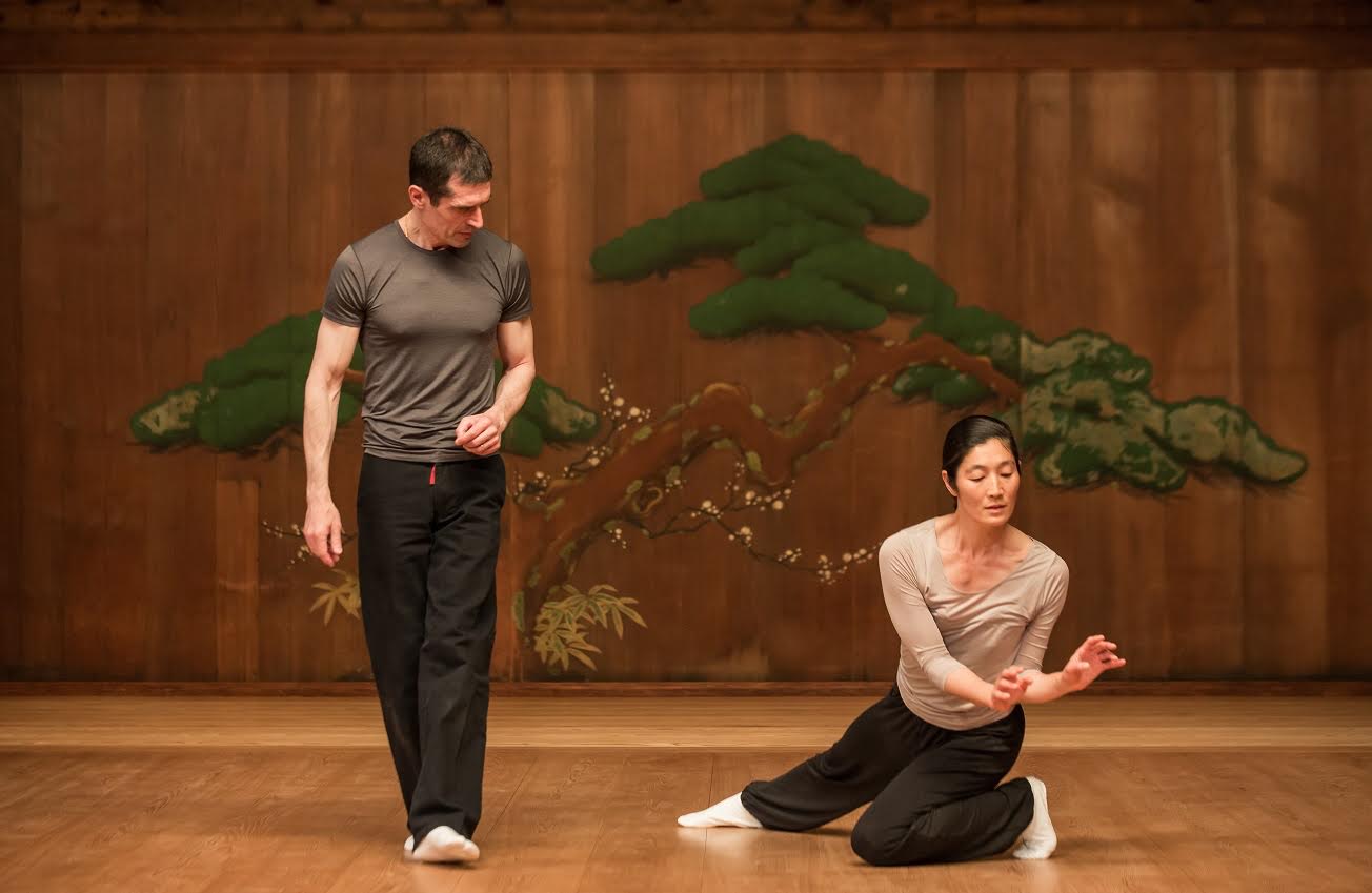 A man watches a woman gesturing on the floor. They both wear black pants and beige tops.