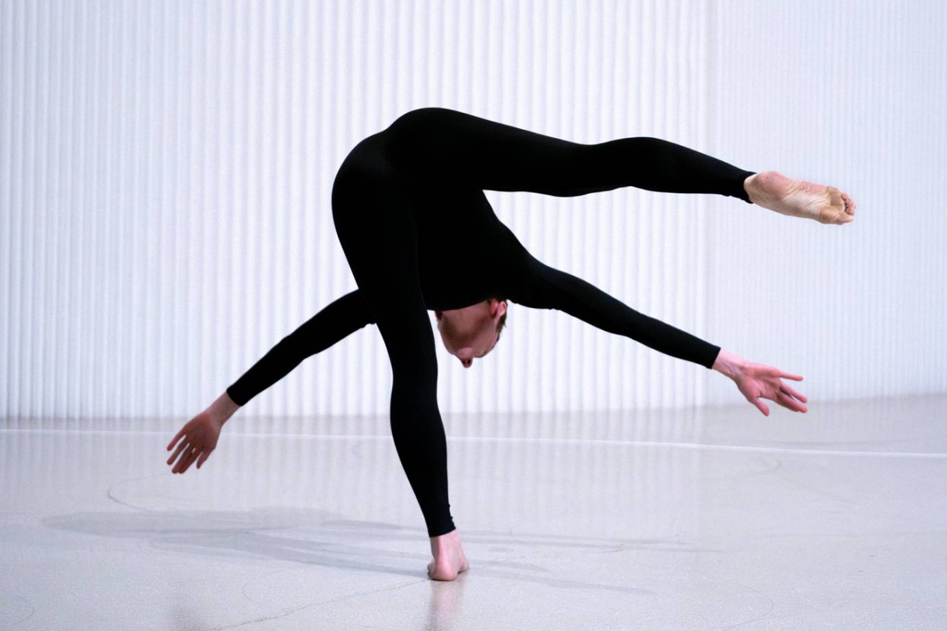 A woman costumed in black balancing on one leg bends deeply forward with her other leg extended behind. 