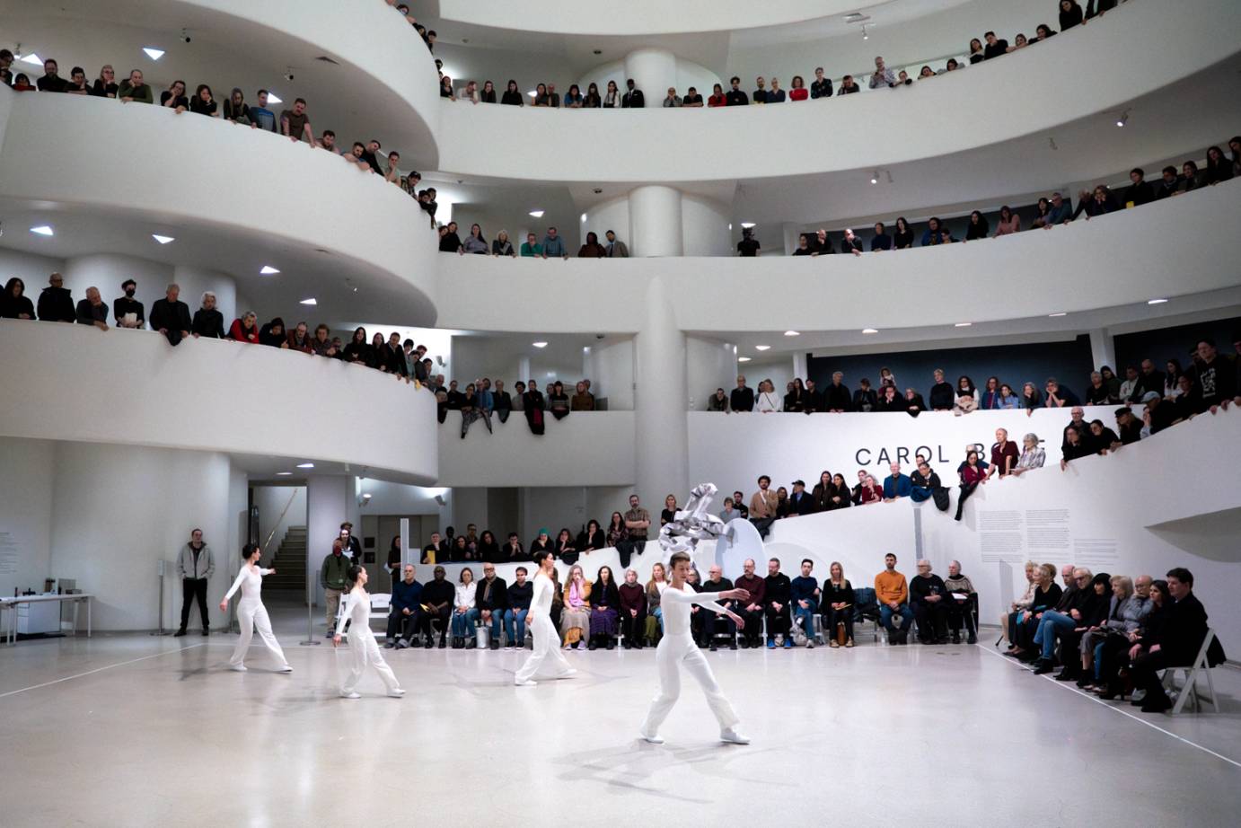 Pictured are three rings of the Guggenheim Museum with audience members standing along the rim. They are watching the four white-costumed dancers performing in the rotunda.