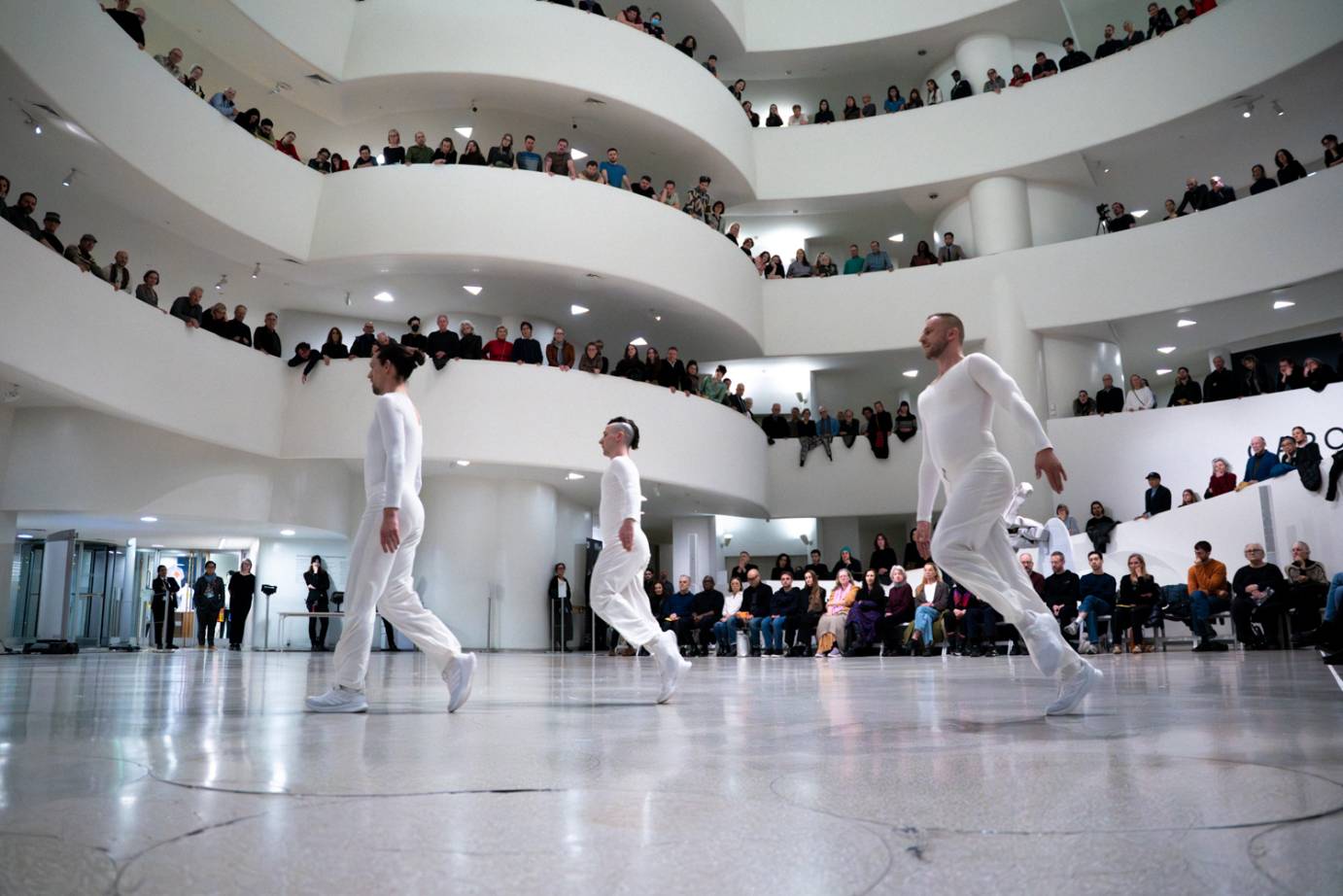 Three of featured four men are in mid-step facing the same direction with the audience draped over the Guggenheim spirals intently watching.