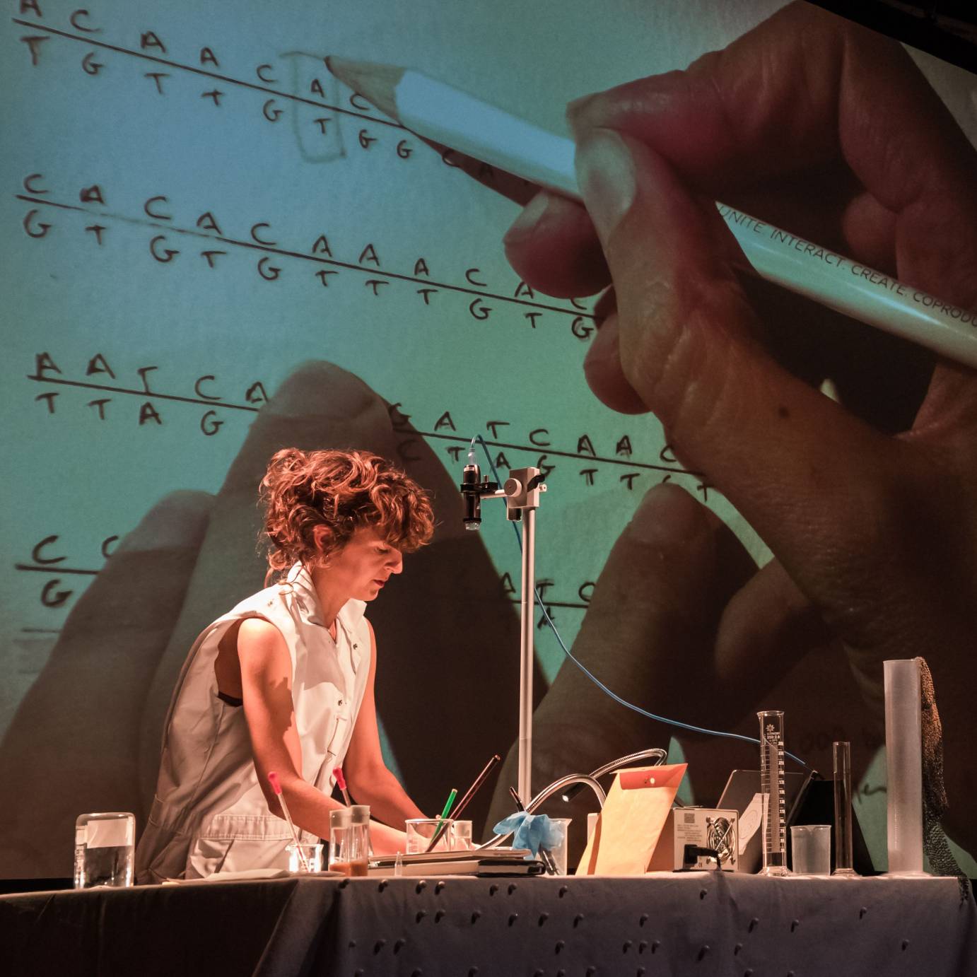 A red-headed woman wearing a white sleeveless shirt sits at a table covered with scientific-looking notebooks, glass bottles, beakers and instruments. 