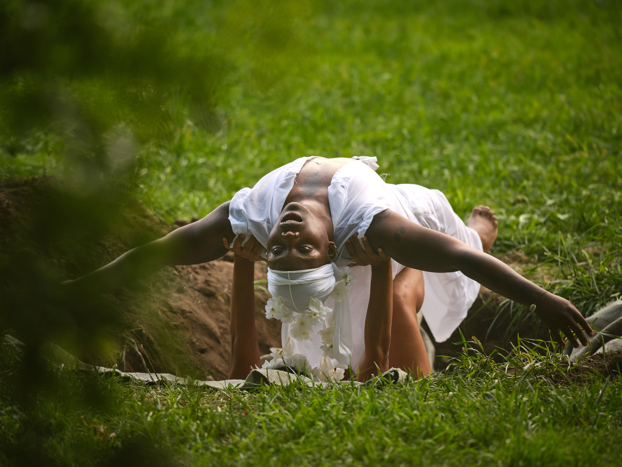 Outdoors, a dancer in a white outfit and du-rag arches dramatically backward over another person's hands and shins. Their arms stretch wide, head tilted upside down, eyes open and gazing directly at the viewer. They are positioned over a patch of dug earth, while the person beneath supports them from within the dug hole. Vibrant green grass surrounds the area.