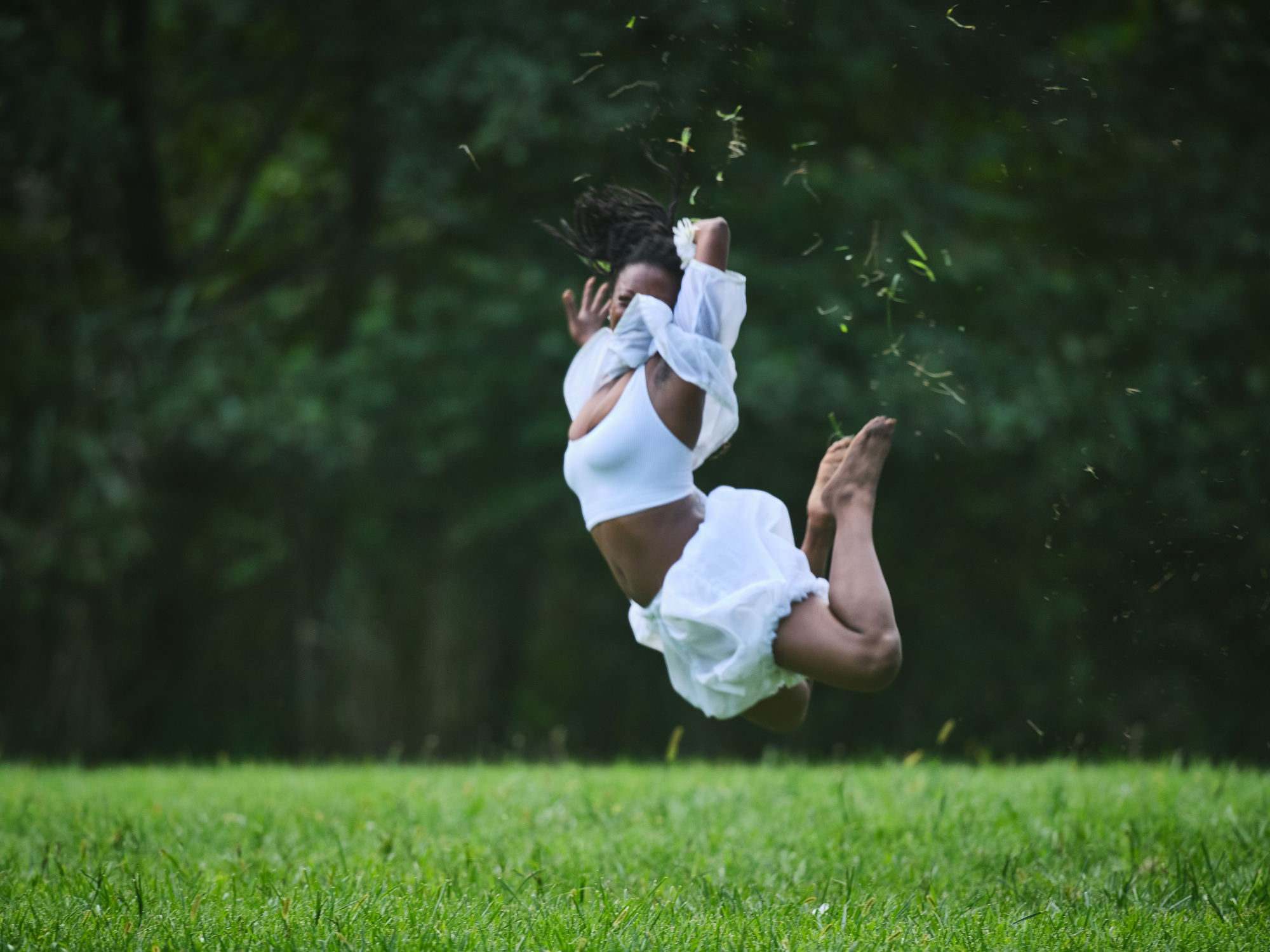  Outdoors, a dancer is caught mid-air in a powerful leap, legs tucked and arms folded near their face. They wear a white cropped shirt and billowy white shorts, with grass and dirt flying around them. The deep green blur of the forest in the background contrasts with the crisp brightness of the dancer’s white costume