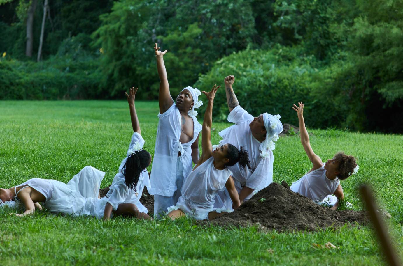   Outdoors on vibrant green grass, a group of six dancers in flowing white garments are arranged in a dynamic pose in and around a mound of earth. They extend one arm skyward while the other remains in contact with the dirt. They wear white flowers in their hair and soft, translucent fabrics, with a backdrop of dense green foliage