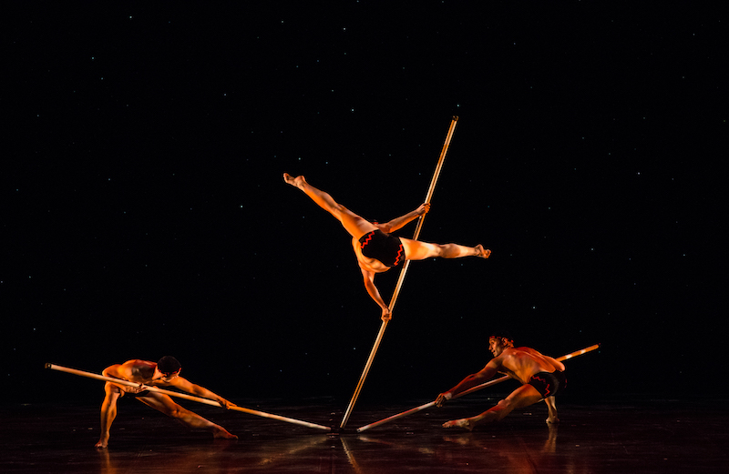 Three men holding long sticks. One man is holding himself in the air by leveraging himself with his pole. 