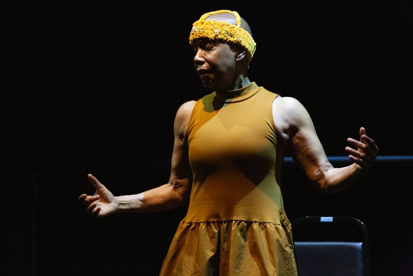 A woman dressed in a mustard shift wearing a mustard-colored hat gestures with both arms on either side of her body.