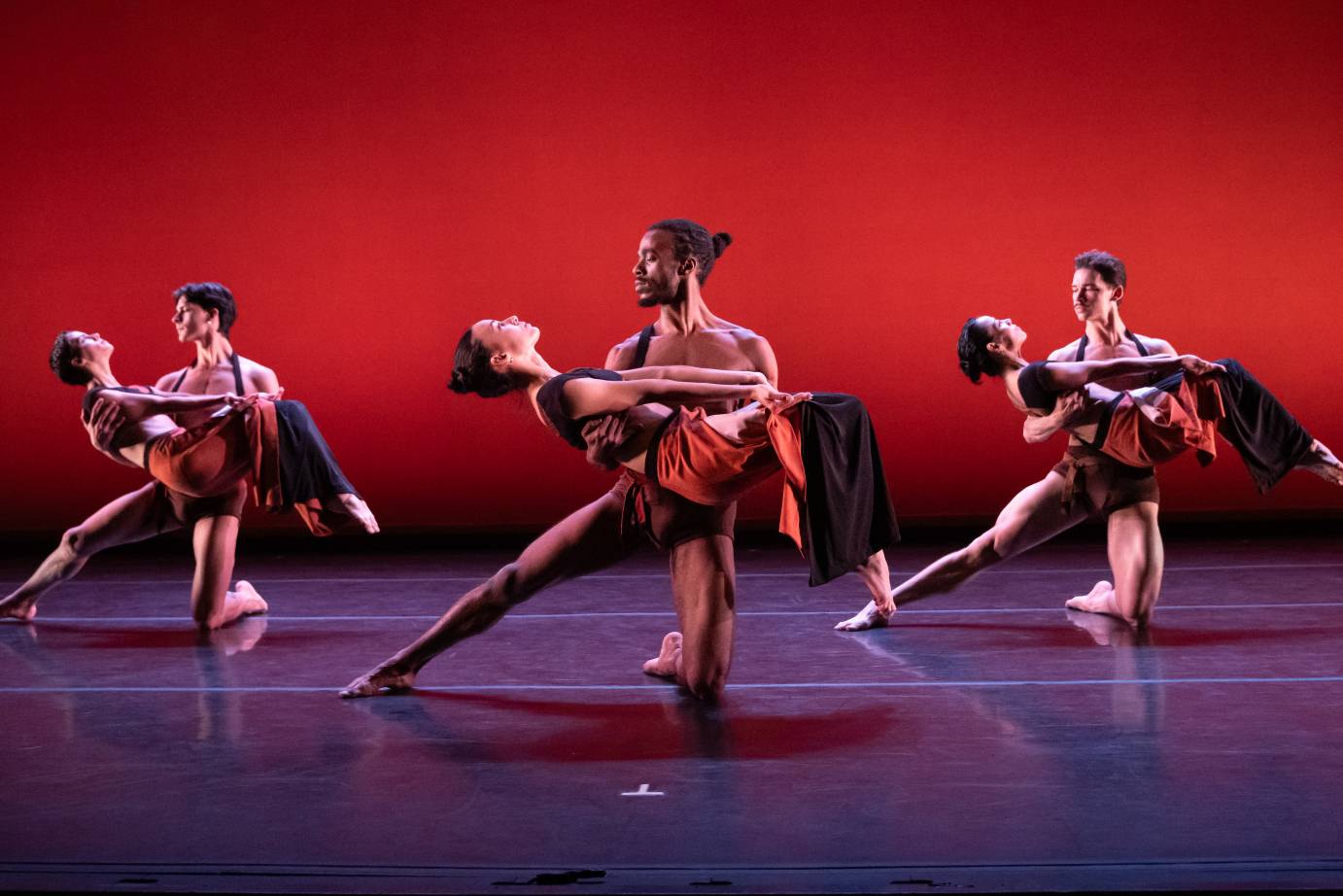 A seated man holds the calves of a woman standing in front of him. She leans forward with one arm extended on the diagonal before her.