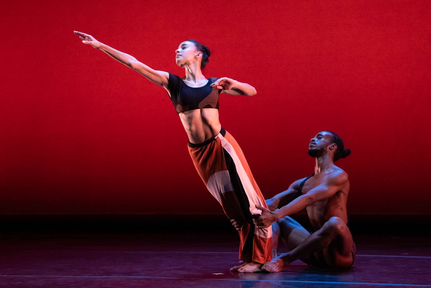 A seated man holds the calves of a standing woman as she leans far forward on the diagonal her right arm extended in front.