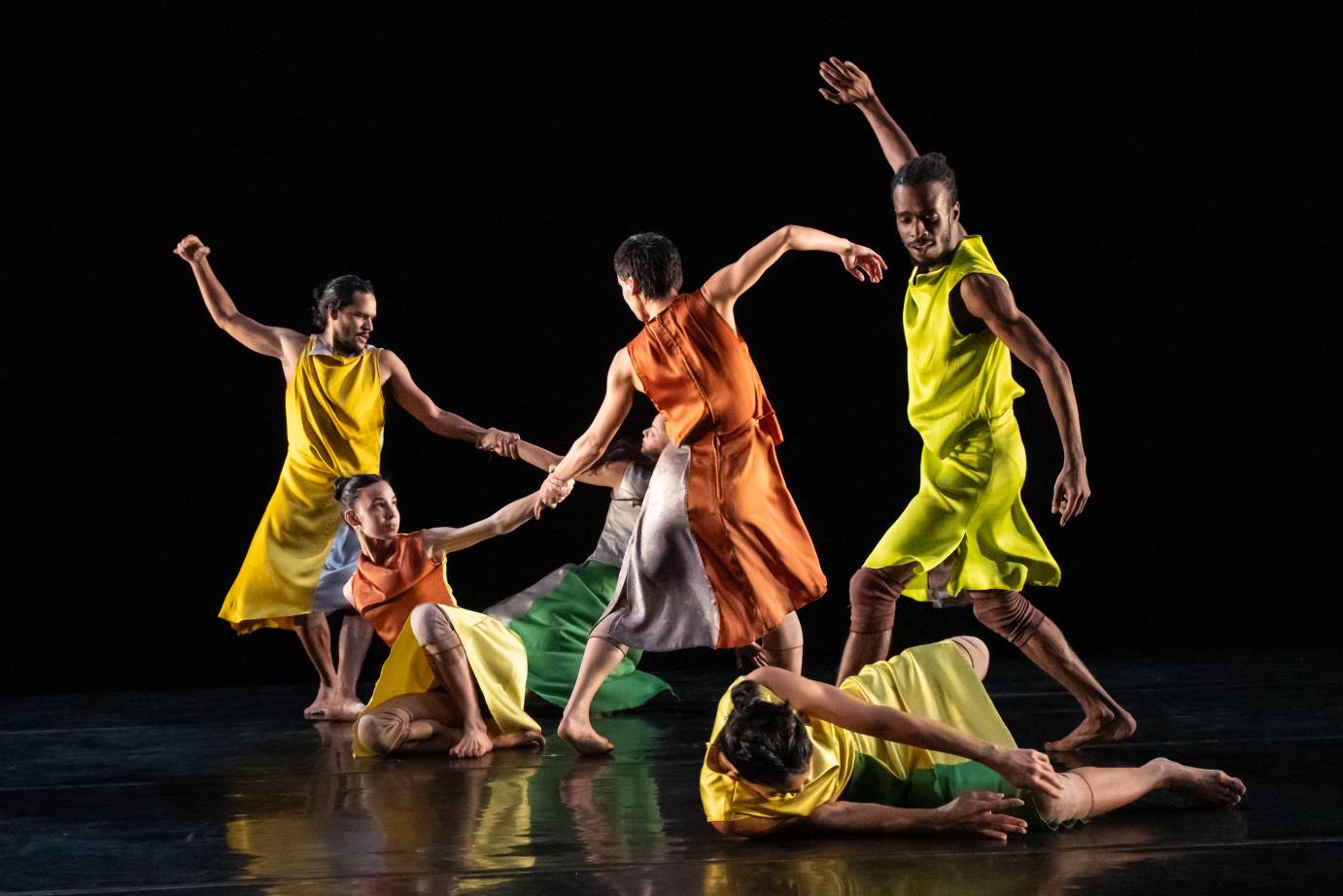 Three dancers in bright skirts and tops move in a circle with one arm flung upward while three are on the ground underfoot.
