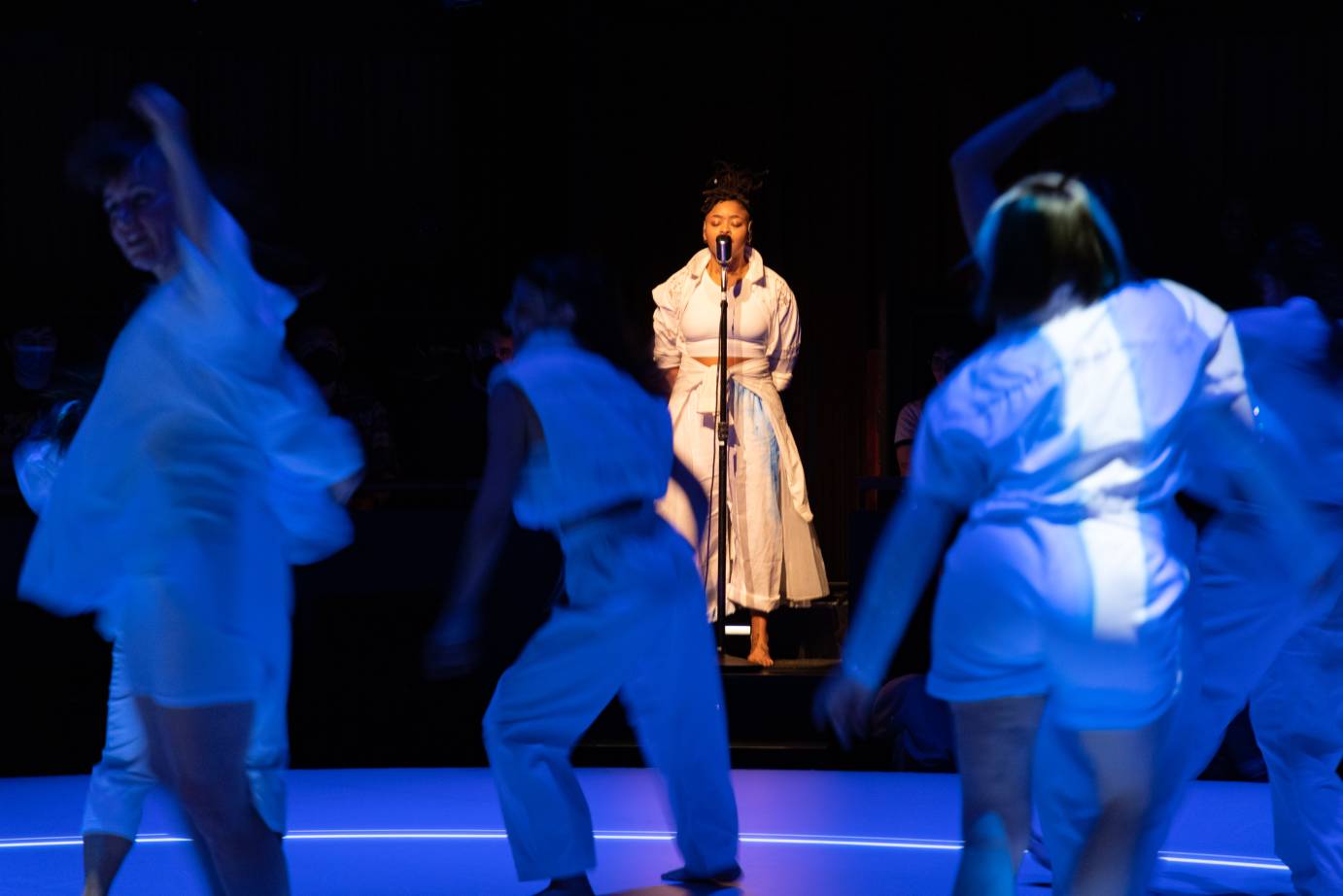 a woman in white outfit speaks into a mike with her eyes closed she speaks in front of a group of women dancing in sheer white costumes.