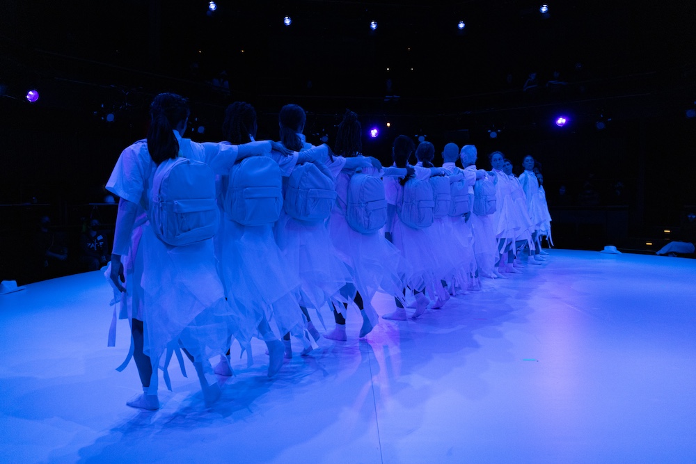 the women in white form a long line across the stage, half of them face one way, the other half the otherway. I notice they have white back packs on their backs.