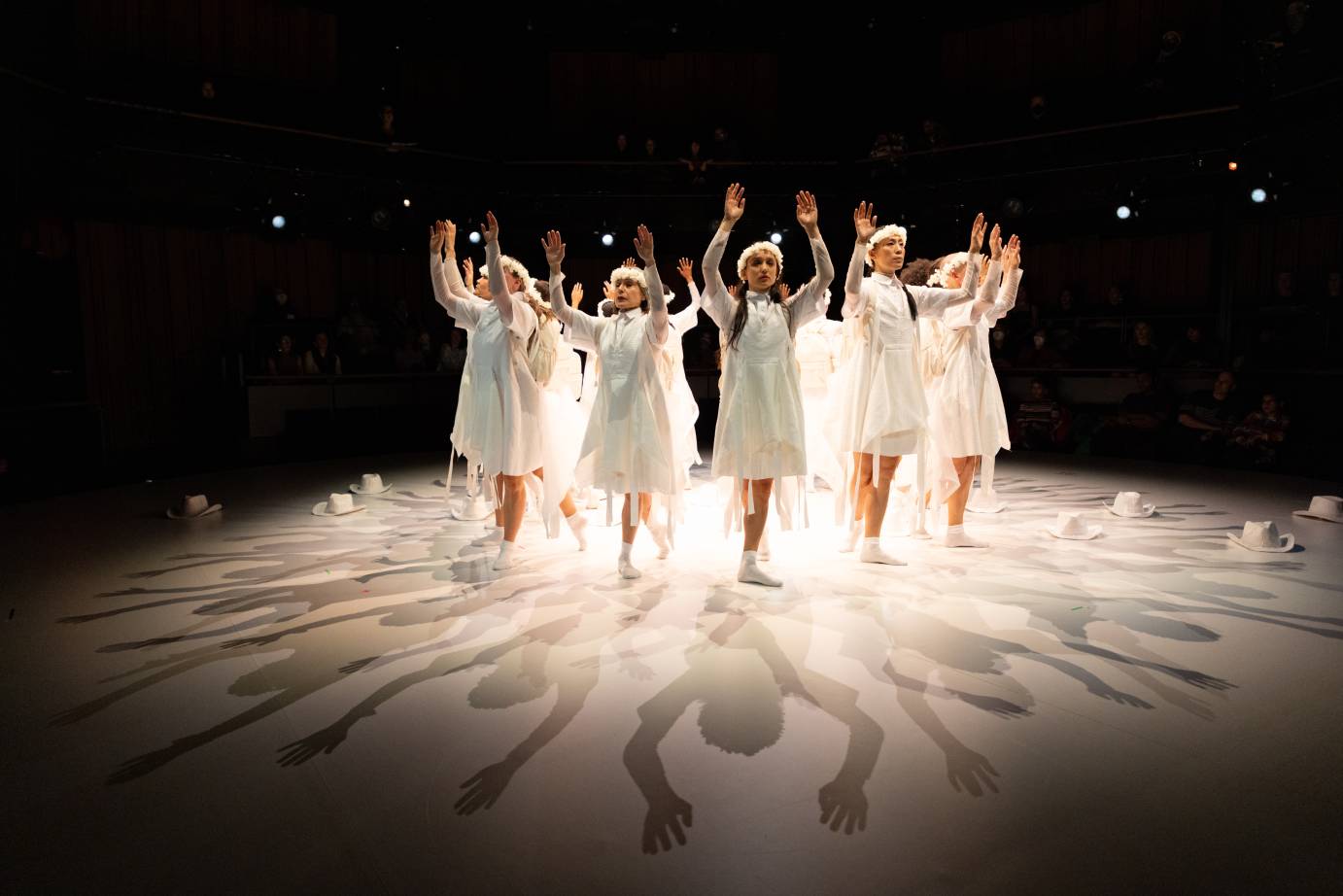 a group of women in a circle facing outward, they wear white filmy dresses and white crowns of what seem to be flowers on their heads, in this picture they raise their hands upward and look almost holy as they do so.