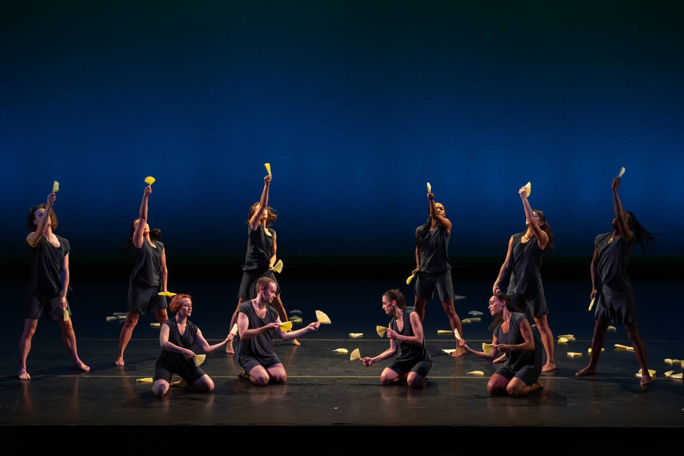 Ten dancers all holding small fans in both hands either kneel and face one another or stand with arms reaching to the sky.