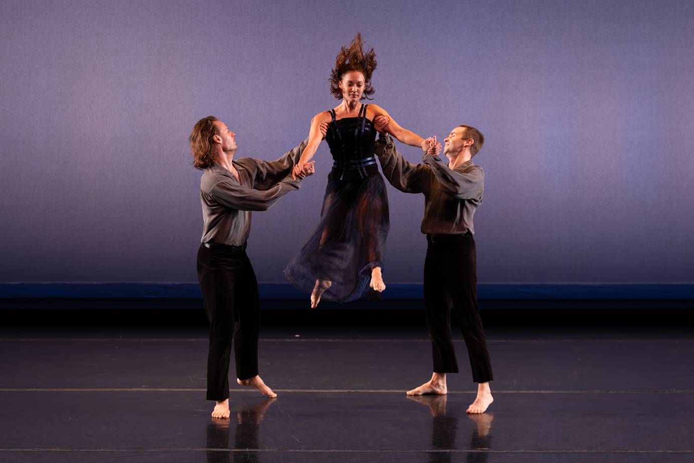 A woman dressed in a sleeveless black dress and hair flying is tossed forward downstage by two men on either side of her.