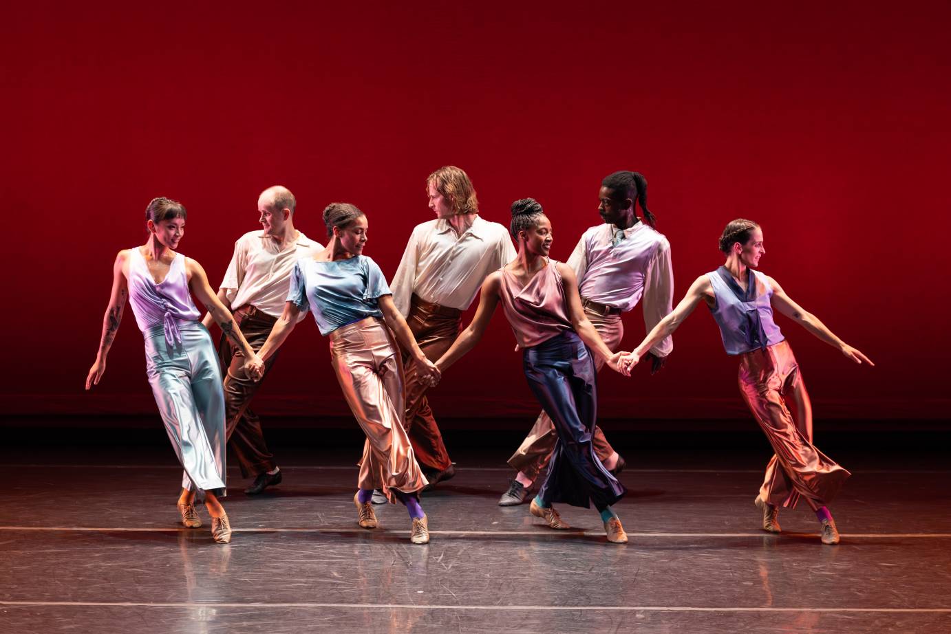 Two rows of dancers - four women in front with their bodies moving to their left and three men behind them move to their right - are dressed in shimmery pastel tops and long pants.