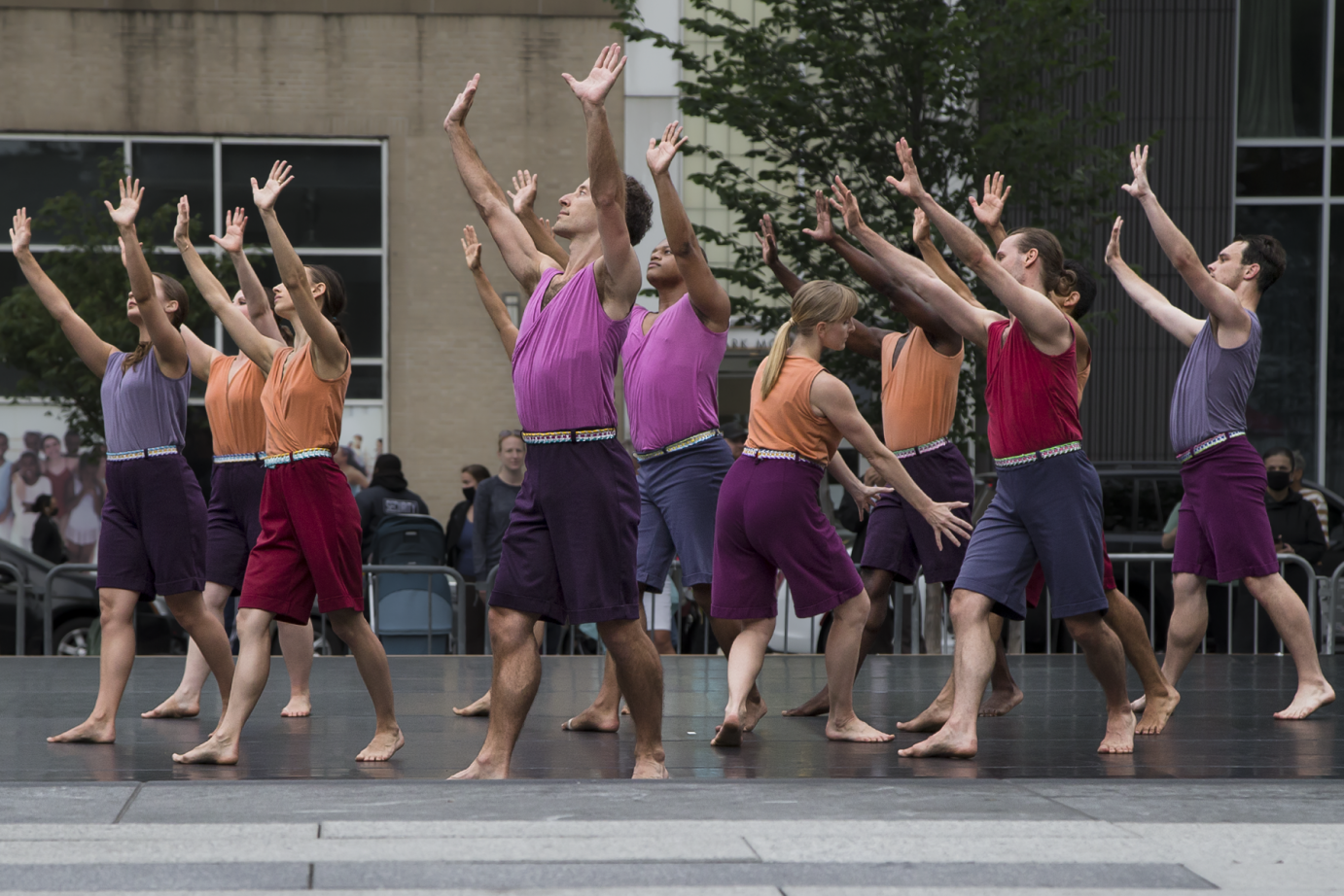 about a dozen dancers of the Mark Morris Dance Group extend their arms on a high diagonal in front of them, almost seeming to be in an act of praise. one dancer looks the other way and her arms reach to the ground 