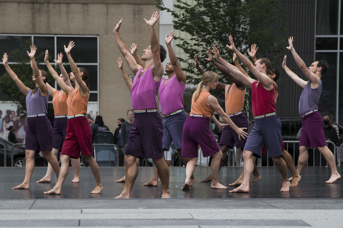 about a dozen dancers of the Mark Morris Dance Group extend their arms on a high diagonal in front of them, almost seeming to be in an act of praise. one dancer looks the other way and her arms reach to the ground