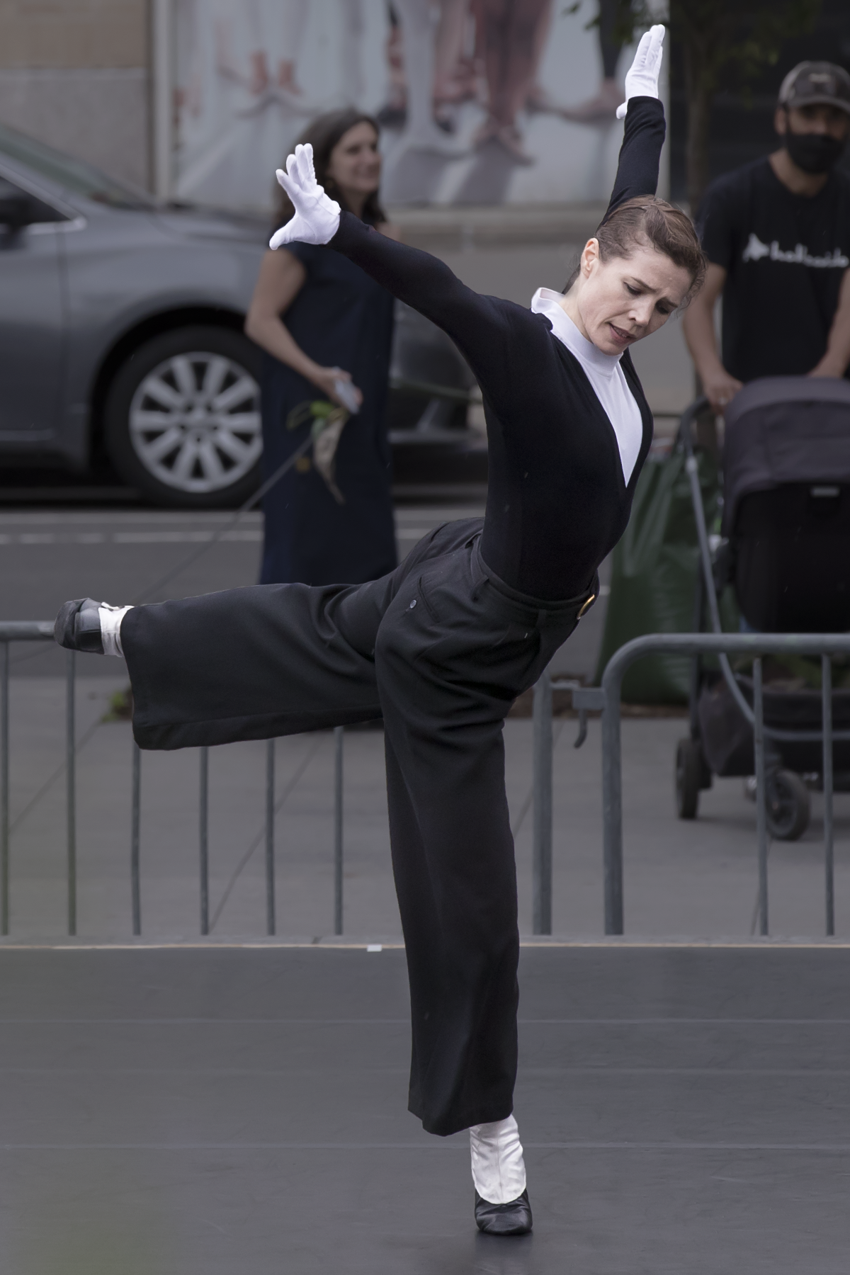 a female dancer with light colored skin wears a black pantsuit with white socks gloves and a bit of white in front of her chest. Her body is fully extended as she balances on one leg with her arms to her sides on high diagnals. She is very concentrated on the music. Her eyes are closed.