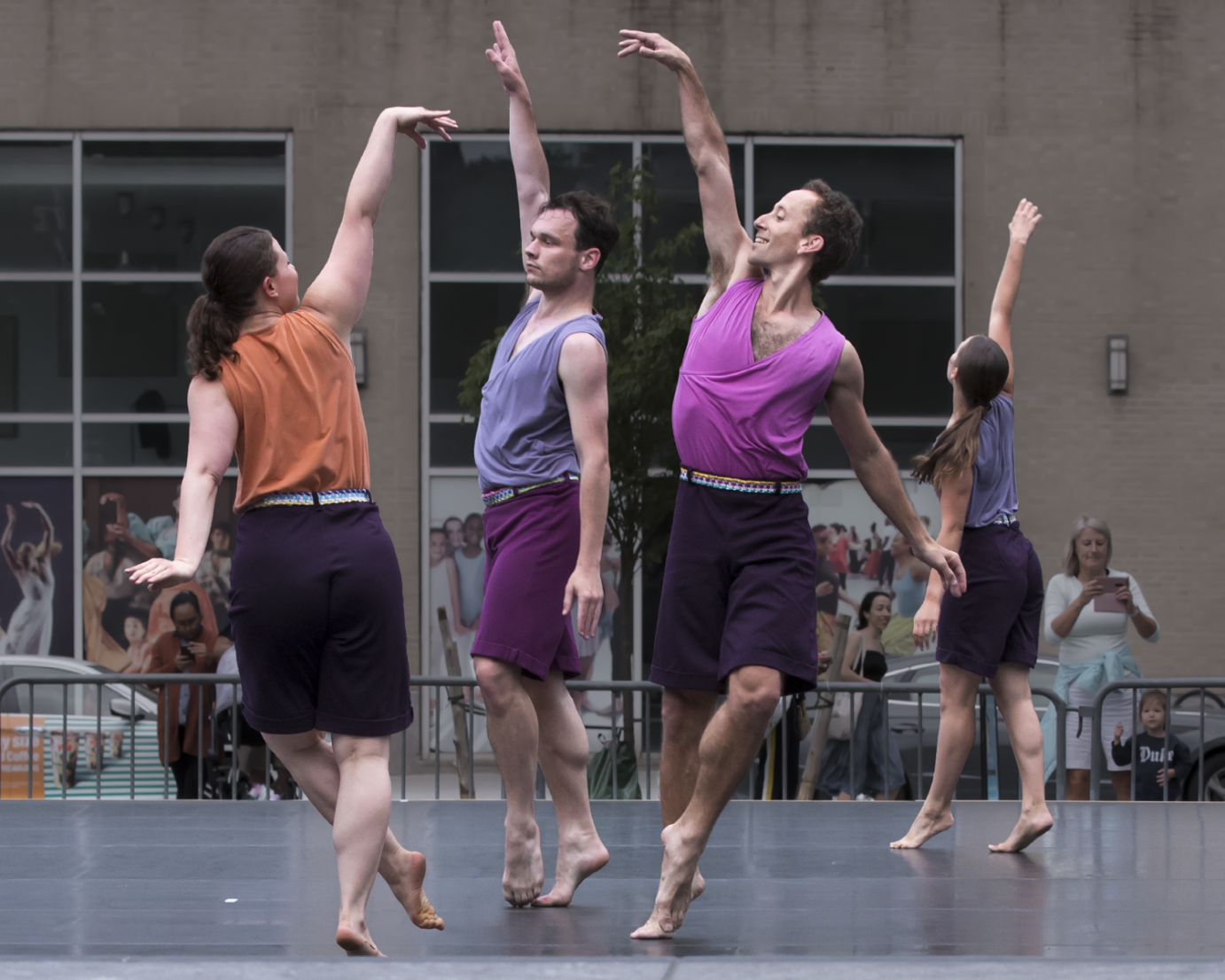 Four dancers two men and two women wearing muted color shorts and sleevles tops look as if they are conversing in a folk dance form their right arms extended into the air