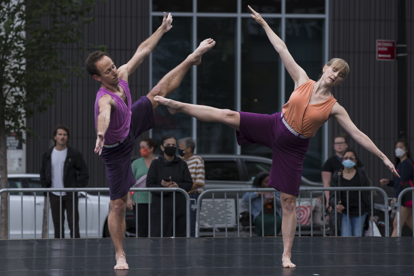 a man and woman dancer both in purple shorts and sleeveless tops stand on one leg tilting to the side with their free leg aiming high into the air