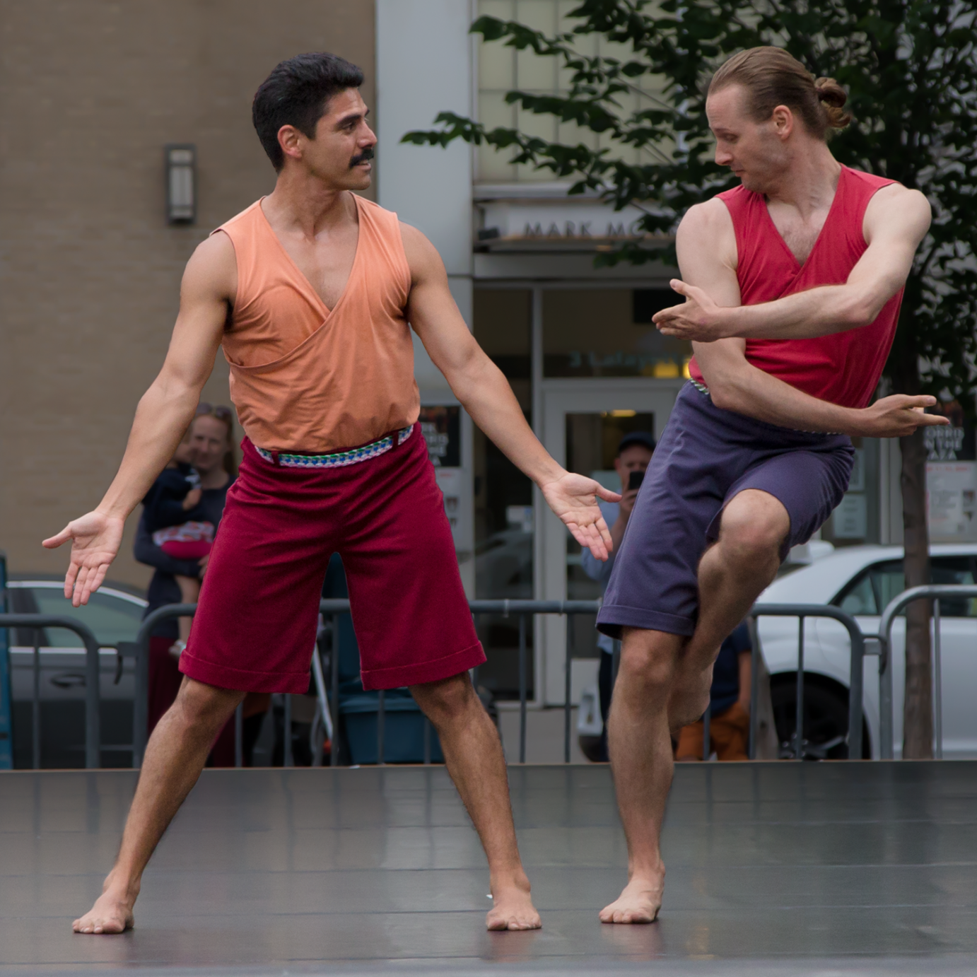 a light tanned skinned dancer with black hair and a moustache looks out at his friend in the middle of  a movement where his arms are wrapped around himself.