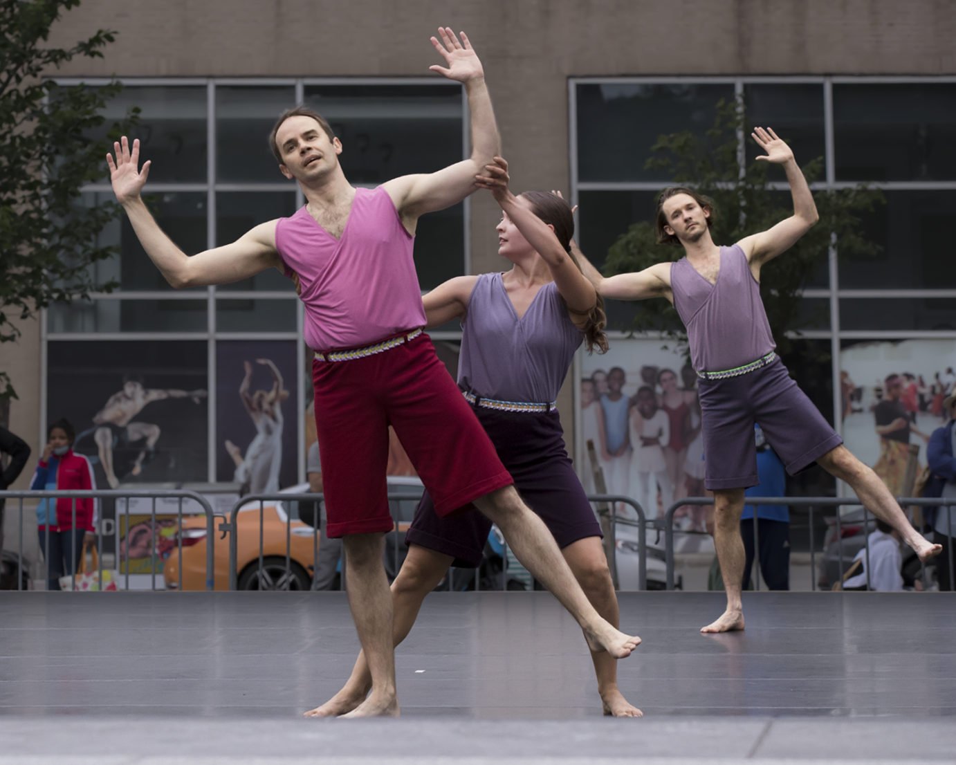 Three dancers in magenta or purple shorts and harmonizing colored sleevles tops are present . One man in the front tilting to the side balancing on one leg as a woman seems to support him and another man behind them replicatiing the first man's pose. 