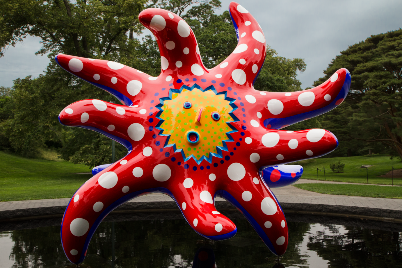 Yayio Kasumas sun sculpture... a yellow dotted face with red rays extending out from it. the whimsical dotted sculpture stands in the middle of a pond surrounded by green grass and tall trees