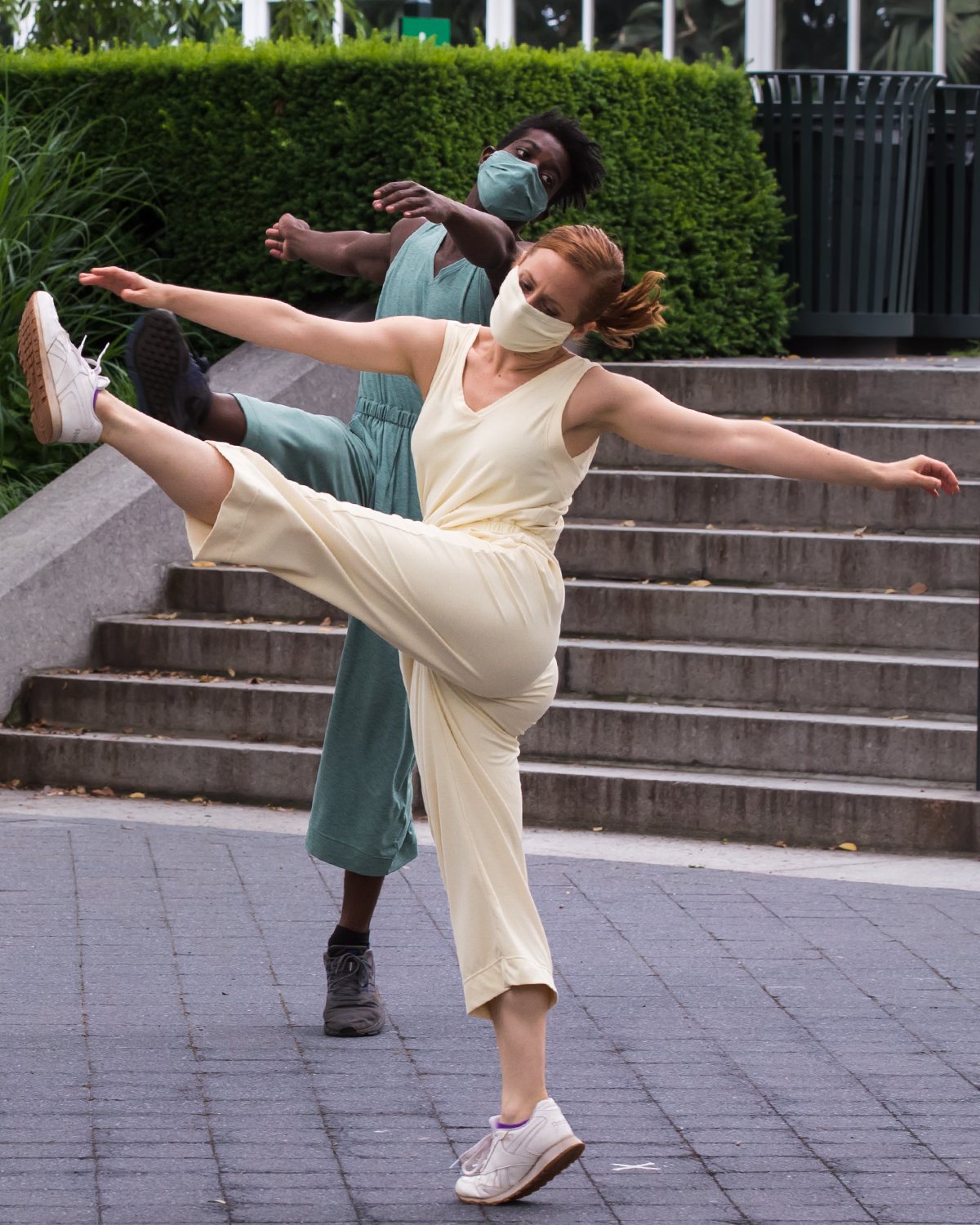 two dancers in pantsuits ... one a seaform green and one a pastel yellow kick one leg high in the air in the middle of a dance, they sport masks that match their costumes.